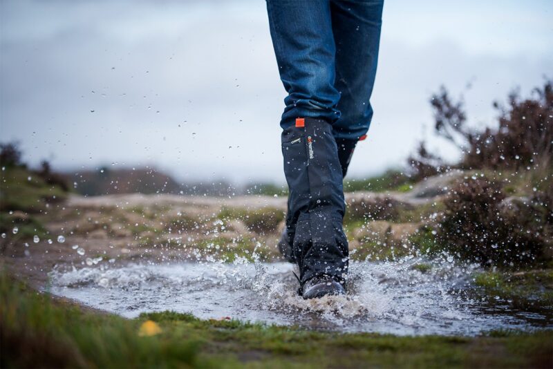 Lower legs in blue jeans and black waterproof gaiters and boots splashing through a muddy puddle on a windswept moorland path, water droplets frozen mid‑air against an overcast sky.