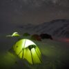 Glowing yellow-green tent in the snowy foreground with several other tents pitched on a snow-covered mountain slope beneath a clear, star-filled night sky and distant peaks.