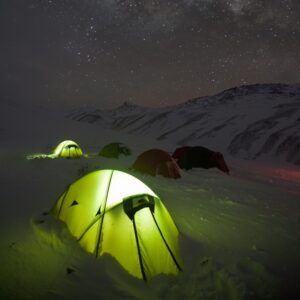 Glowing yellow-green tent in the snowy foreground with several other tents pitched on a snow-covered mountain slope beneath a clear, star-filled night sky and distant peaks.