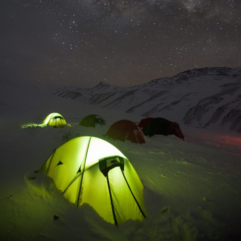 Glowing yellow-green tent in the snowy foreground with several other tents pitched on a snow-covered mountain slope beneath a clear, star-filled night sky and distant peaks.