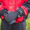 Person wearing a red-and-black jacket holding padded black mittens with a strap and visible "extremities" logo against a blurred outdoor background.