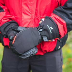Person wearing a red-and-black jacket holding padded black mittens with a strap and visible "extremities" logo against a blurred outdoor background.