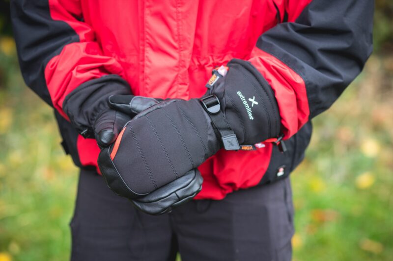 Person wearing a red-and-black jacket holding padded black mittens with a strap and visible "extremities" logo against a blurred outdoor background.