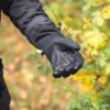 Close-up of a person in a black jacket holding out a black insulated glove palm-up to show its textured leather palm, with blurred yellow autumn leaves in the background.