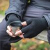 Hands in black fingerless gloves holding a stainless-steel cup while sitting on an outdoor bench, wearing a dark jacket with blurred autumn foliage in the background.