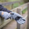 Hands wearing grey wool gloves with black palm patches and an orange 'XDRY' label resting on a weathered wooden railing, the wearer in a dark jacket on an outdoor boardwalk.