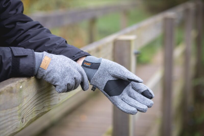 Hands wearing grey wool gloves with black palm patches and an orange 'XDRY' label resting on a weathered wooden railing, the wearer in a dark jacket on an outdoor boardwalk.