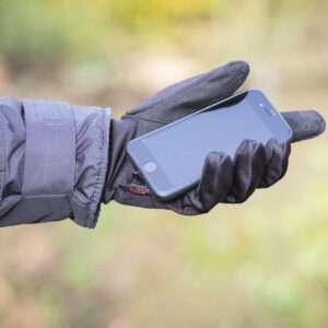 A person in a black winter jacket and touchscreen-compatible black glove holds a black smartphone in their outstretched hand against a soft-focused green and brown outdoor background.