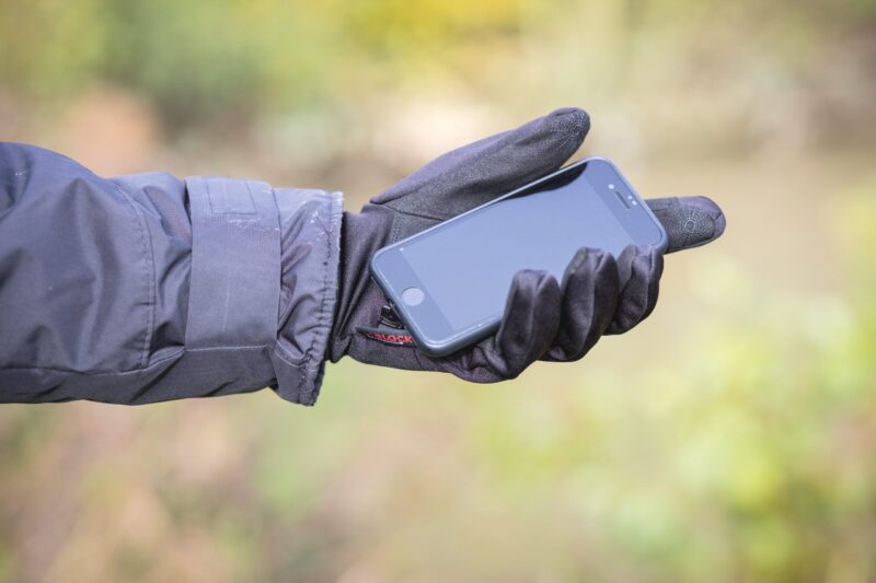 A person in a black winter jacket and touchscreen-compatible black glove holds a black smartphone in their outstretched hand against a soft-focused green and brown outdoor background.