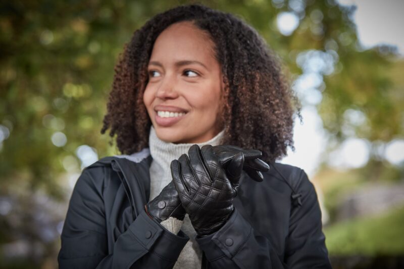 Woman with curly hair outdoors, smiling and looking to the left while clasping quilted black leather gloves and wearing a dark jacket over a light turtleneck.