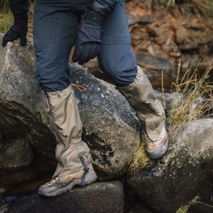 Lower legs of a hiker wearing beige waterproof gaiters over muddy brown hiking boots and dark trousers, stepping across wet river rocks beside grassy banks.