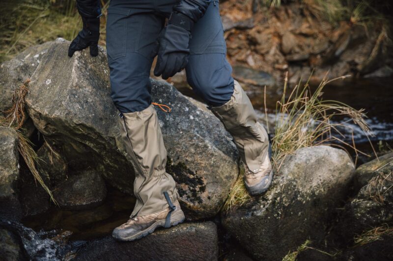 Lower legs of a hiker wearing beige waterproof gaiters over muddy brown hiking boots and dark trousers, stepping across wet river rocks beside grassy banks.