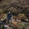 A man in a dark puffer jacket, gloves and gaiters sits on a rock beside a shallow stream, looking upward amid ferns, mossy stones and a dense heather-covered hillside.