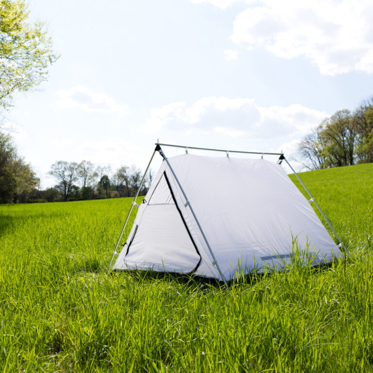 White A-frame tent with metal poles pitched in a sunlit green meadow, triangular zipped door visible and a tree line and blue sky with scattered clouds in the background.