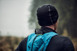Flux-Beanie-5-scaled-2.jpg Person seen from behind wearing a black beanie and jacket with a bright blue backpack, standing in a misty, blurred forest background.