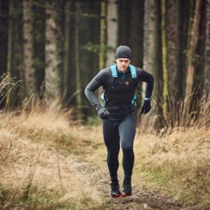 A runner wearing black leggings, a black long-sleeve top, gloves and a beanie with a blue hydration backpack, running on a narrow forest path lined with dry grasses and tall conifer trunks.