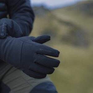 Close-up of a person adjusting dark insulated Extremities gloves with a blurred grassy hillside in the background.