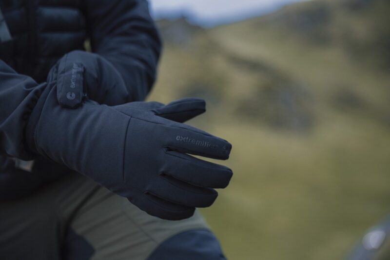 Close-up of a person adjusting dark insulated Extremities gloves with a blurred grassy hillside in the background.