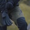 Close-up of a person wearing black insulated gloves and olive-green outdoor trousers with reinforced black knee panels and a zipped thigh pocket, hands extended against a blurred grassy hillside background.