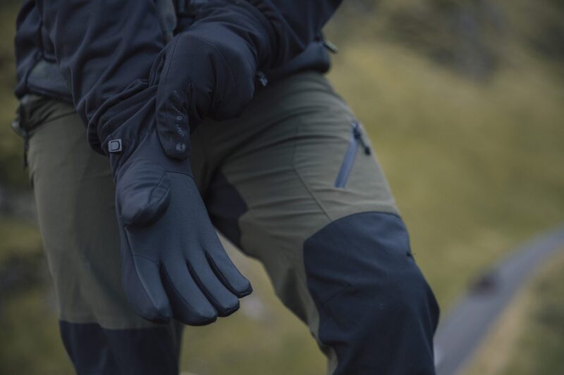 Close-up of a person wearing black insulated gloves and olive-green outdoor trousers with reinforced black knee panels and a zipped thigh pocket, hands extended against a blurred grassy hillside background.