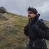A man in a black puffer jacket and gloves carrying a backpack hikes across grassy rolling hills under an overcast sky with a solitary tree on a distant ridge.