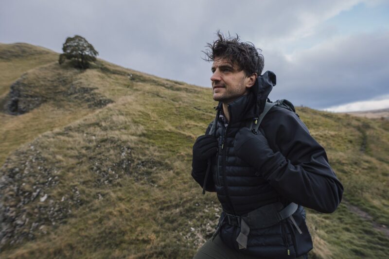 A man in a black puffer jacket and gloves carrying a backpack hikes across grassy rolling hills under an overcast sky with a solitary tree on a distant ridge.