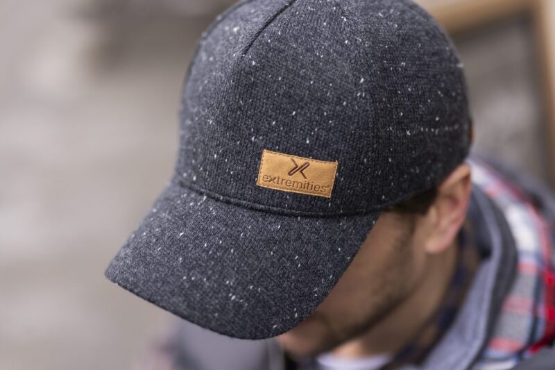 Dark grey flecked baseball cap with a tan leather 'extremities' patch, worn tilted forward on a man's head with his face blurred in the background.