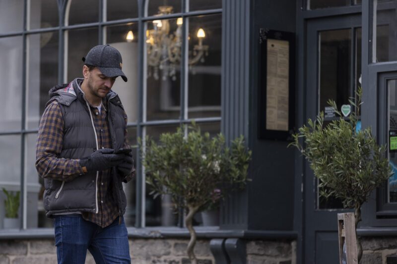 Man in a dark cap, padded vest, checked shirt and gloves walking past a café window while looking at his smartphone, a chandelier visible inside and potted trees by the entrance
