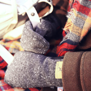 Cozy winter accessories featuring a close-up of a hand wearing a gray knitted glove, surrounded by a colorful plaid scarf and a brown jacket, perfect for cold weather fashion.