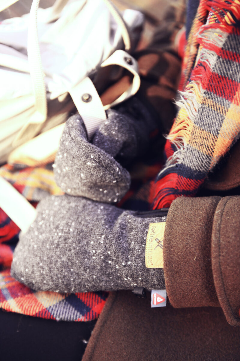 Cozy winter accessories featuring a close-up of a hand wearing a gray knitted glove, surrounded by a colorful plaid scarf and a brown jacket, perfect for cold weather fashion.