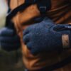 Close-up of hands in speckled dark grey gloves with a small zipped pocket and leather logo patch, gripping backpack straps over an orange outdoor jacket against a blurred woodland background.