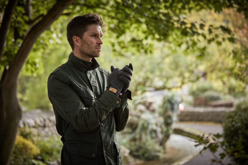 Man in a dark green jacket and grey gloves stands under leafy trees, clasping his hands and looking pensively to the left with a blurred garden path and greenery behind him.
