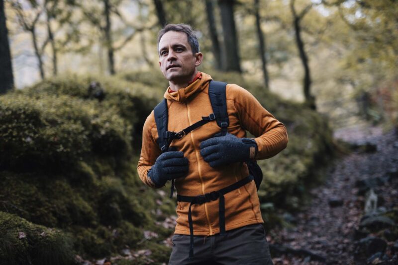 Man in an orange fleece jacket and grey speckled gloves adjusting his backpack's chest strap while walking on a moss-covered woodland path with trees and autumnal foliage.