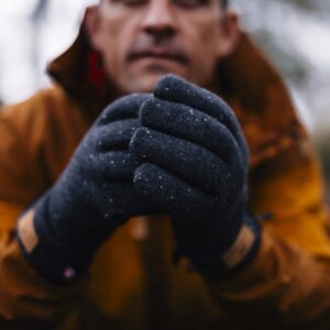 Close-up of a person in an orange jacket holding their dark flecked winter gloves together, with their face blurred in the background.