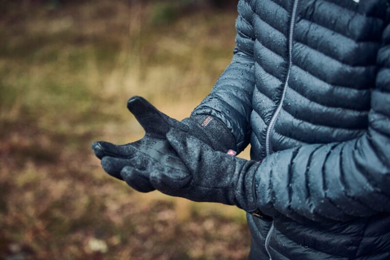 A person in a wet dark blue quilted puffer jacket and black gloves, both dotted with raindrops, holds out cupped hands against a blurred woodland background.