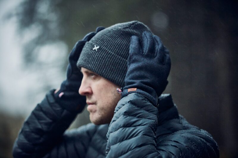 Profile of a man outdoors in light rain adjusting a dark ribbed beanie with a small white X logo while wearing black gloves and a wet quilted puffer jacket against a blurred forest background.