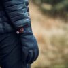 Close-up of a person’s gloved hand and black water-repellent puffer jacket sleeve marked HYDROLOFT, with small fabric tags on the glove cuff against a blurred brown grassy background.