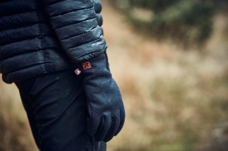 Close-up of a person’s gloved hand and black water-repellent puffer jacket sleeve marked HYDROLOFT, with small fabric tags on the glove cuff against a blurred brown grassy background.