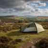 Scenic view of a camping tent set up on a grassy hillside, surrounded by rocky formations and rolling green fields under a cloudy sky, ideal for outdoor enthusiasts and nature lovers.