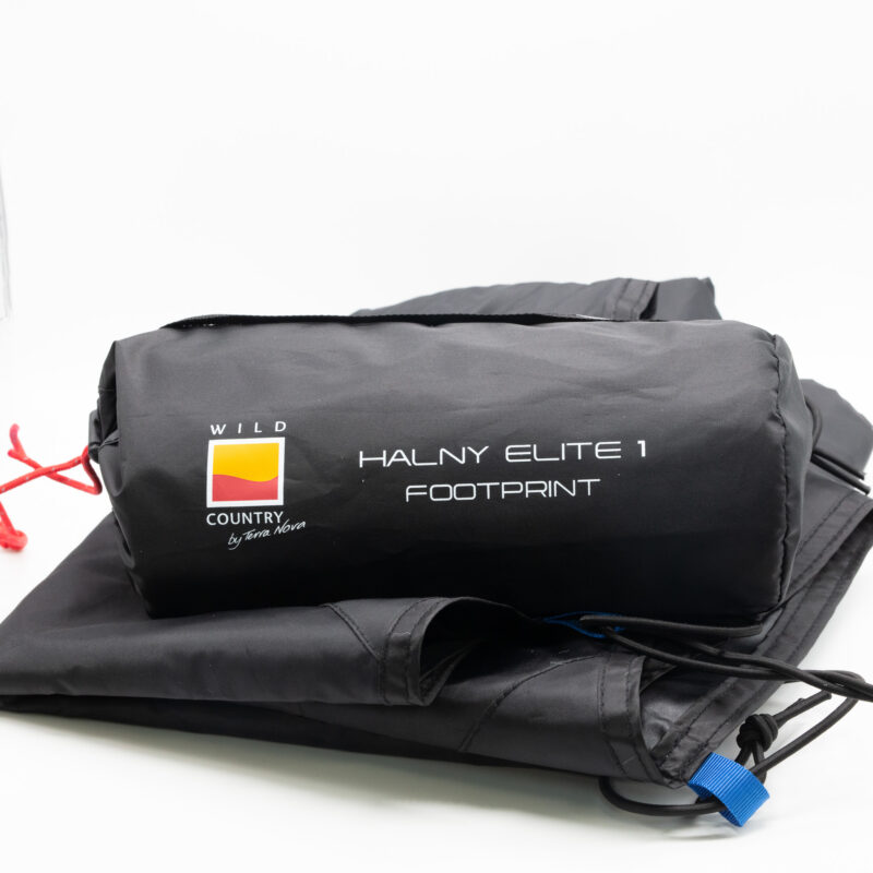 Black rolled tent footprint in its storage bag labeled "HALNY ELITE 1 FOOTPRINT" with a yellow‑red Wild Country logo, red drawcord and blue tab, lying on a folded black groundsheet against a white background.