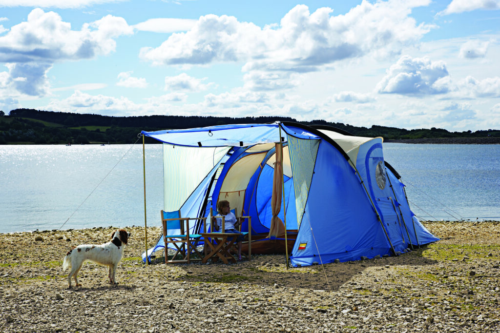 Blue family tent with an awning pitched on a pebbly lakeshore, a child sitting on a folding chair inside, a white-and-brown dog standing nearby and calm water with tree-lined hills under a partly cloudy sky behind them.