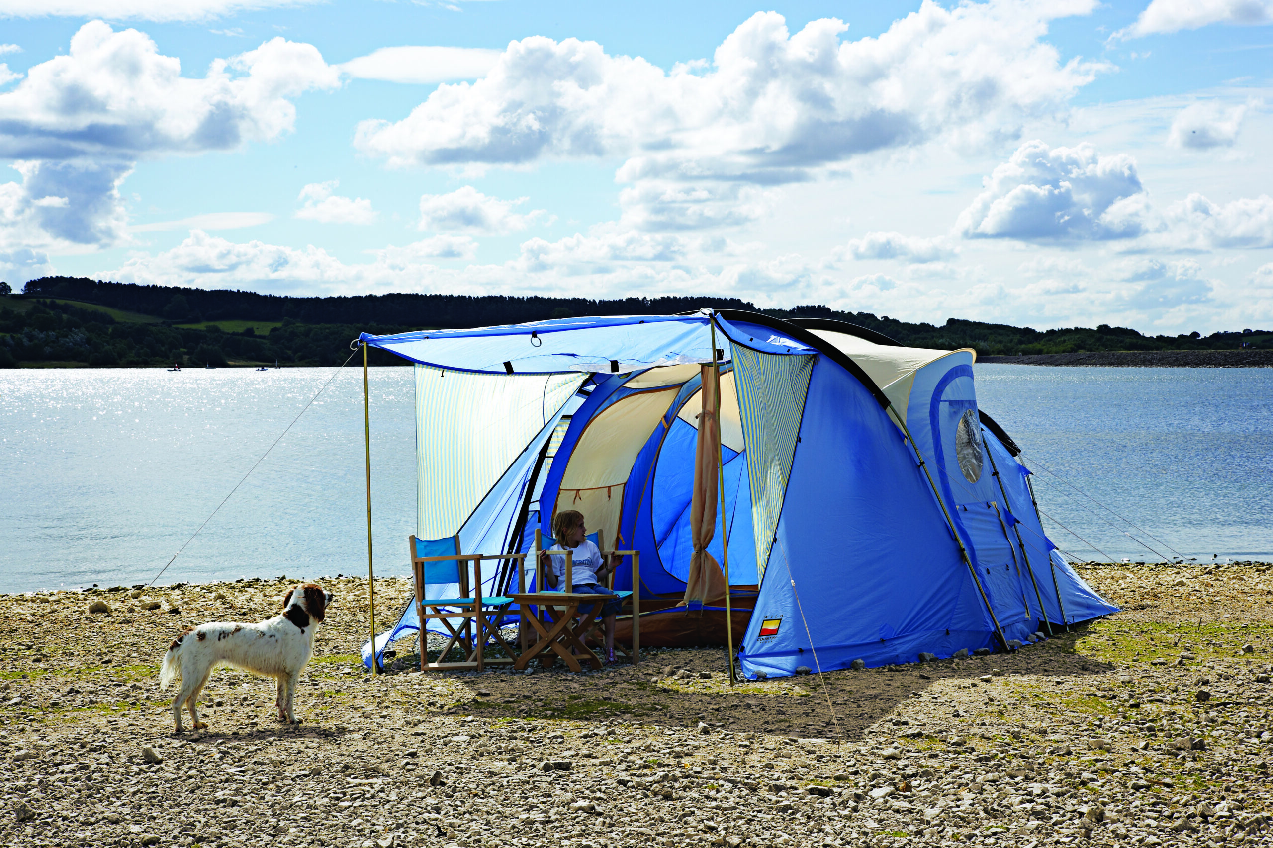 Blue family tent with an awning pitched on a pebbly lakeshore, a child sitting on a folding chair inside, a white-and-brown dog standing nearby and calm water with tree-lined hills under a partly cloudy sky behind them.