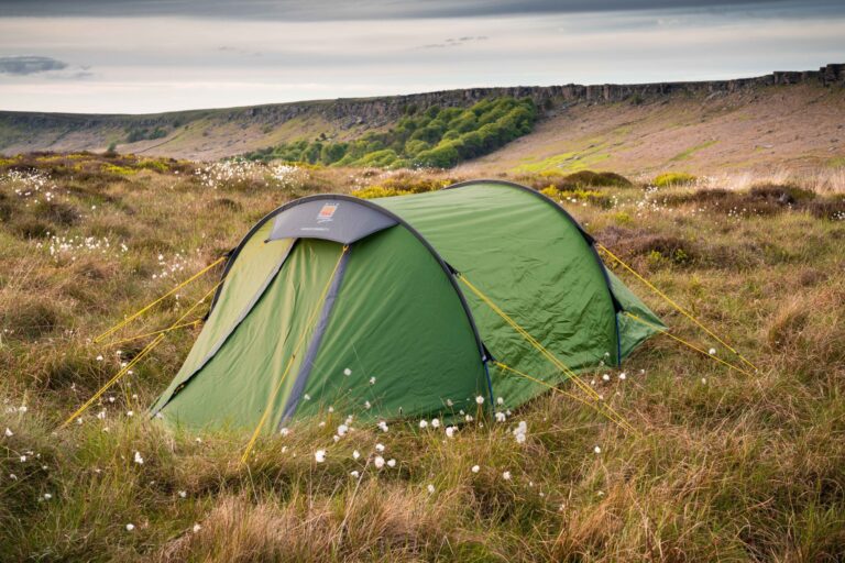 Green tunnel tent secured with bright yellow guy lines pitched among tall grass and white cottongrass on a windswept moor, with a rocky escarpment and a band of trees beneath a cloudy sky in the background.
