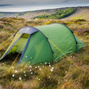 A green tunnel tent pegged with yellow guy lines pitched on windswept heather and cotton-grass moorland with low rocky cliffs and a cloudy sky in the background.