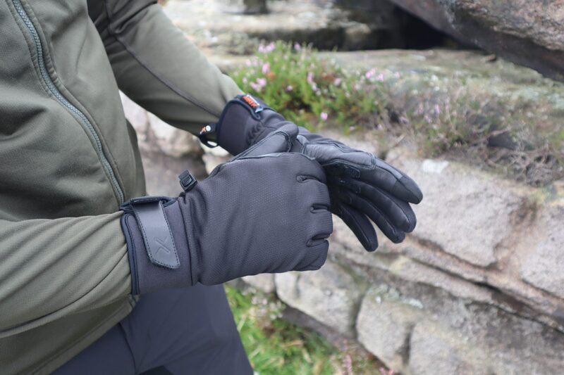 Person in an olive-green jacket putting on black insulated gloves with wrist straps and textured leather palms beside lichen-covered rocks and small pink flowers