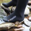 Close-up of a black sock branded 'extremities' on a foot standing on wet river stones with shallow water flowing around them.