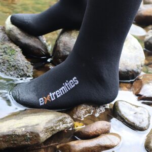 Close-up of a black sock branded 'extremities' on a foot standing on wet river stones with shallow water flowing around them.