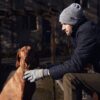 Man in a grey beanie and navy quilted jacket sits on a bench outdoors, reaching with a gloved hand to pet a brown short‑haired dog wearing a rope collar, with an old stone building in the background.