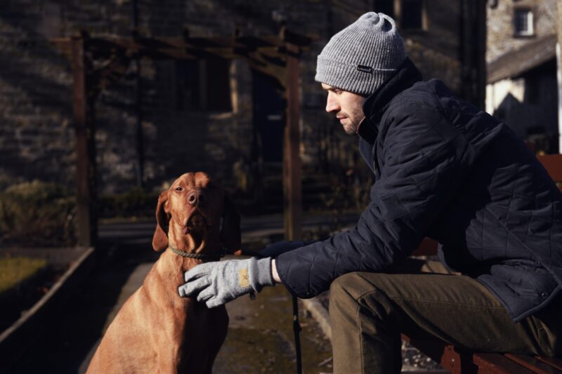 Man in a grey beanie and navy quilted jacket sits on a bench outdoors, reaching with a gloved hand to pet a brown short‑haired dog wearing a rope collar, with an old stone building in the background.