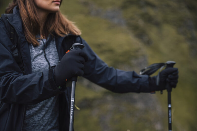 Close-up of a woman wearing gloves and a jacket, holding a hiking pole while exploring a mountainous landscape. The image captures the spirit of outdoor adventure and hiking gear essentials.